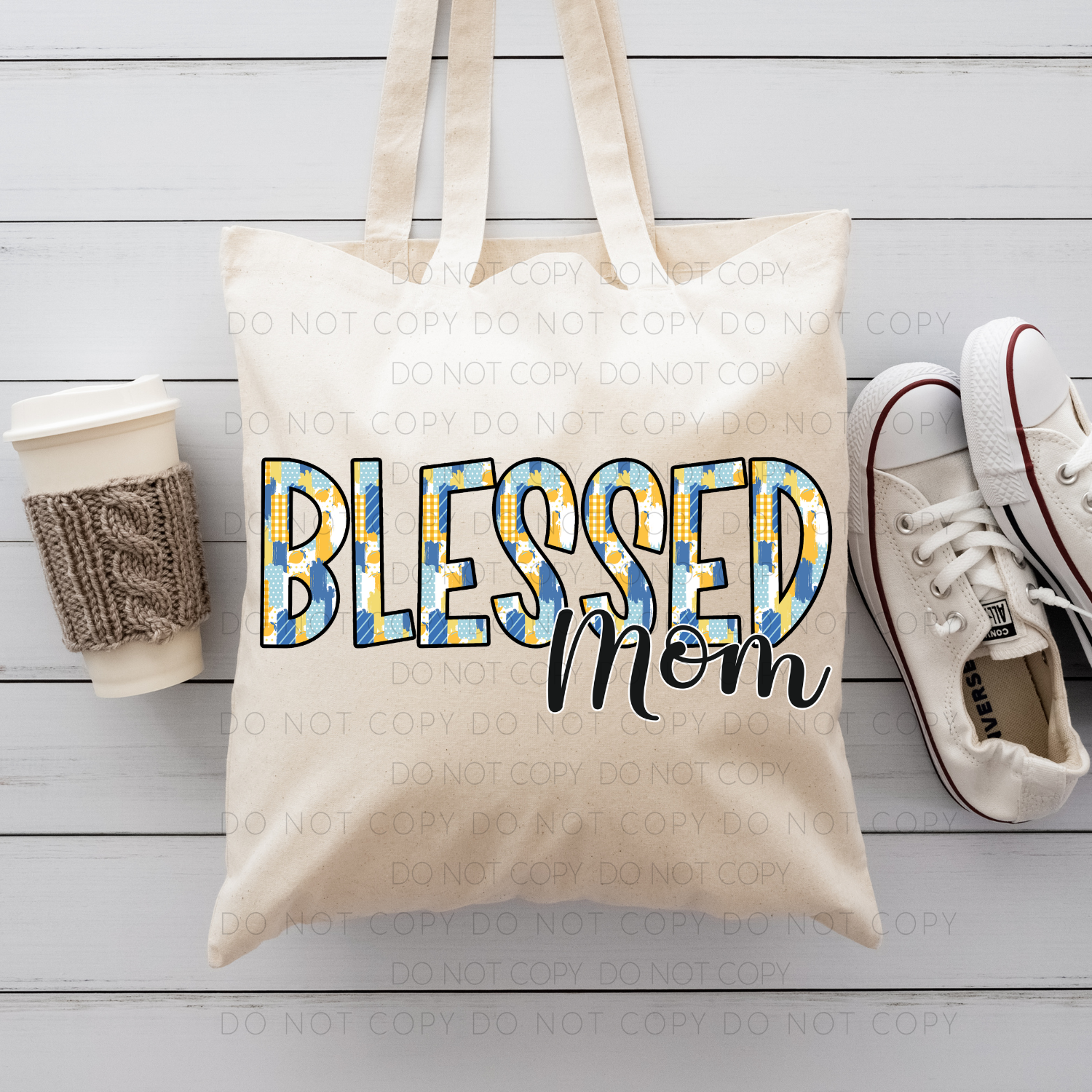 A beige cotton tote bag with 'Blessed Mom' printed in blue and yellow dual-sided DTF print, accompanied by a pair of white sneakers and a brown knit hat on a wooden surface.
