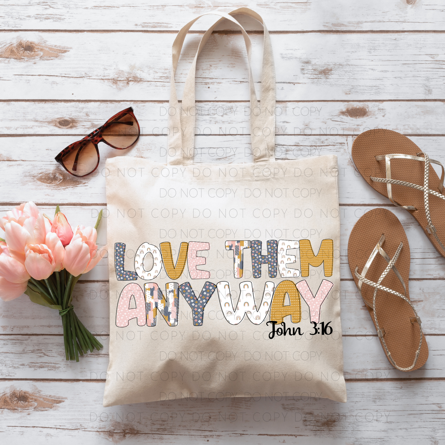 A beige 100% cotton tote bag with a printed phrase 'LOVE THEM ANYWAY' and a Bible reference 'John 16'. The bag is displayed on a wooden surface accompanied by pink tulips, a pair of sunglasses, and a pair of sandals.