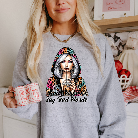 A person wearing a gray unisex t-shirt with a graphic design of a woman holding a coffee cup and text 'Good Women Say Bad Words'. The person is also holding a red and white checkered mug.