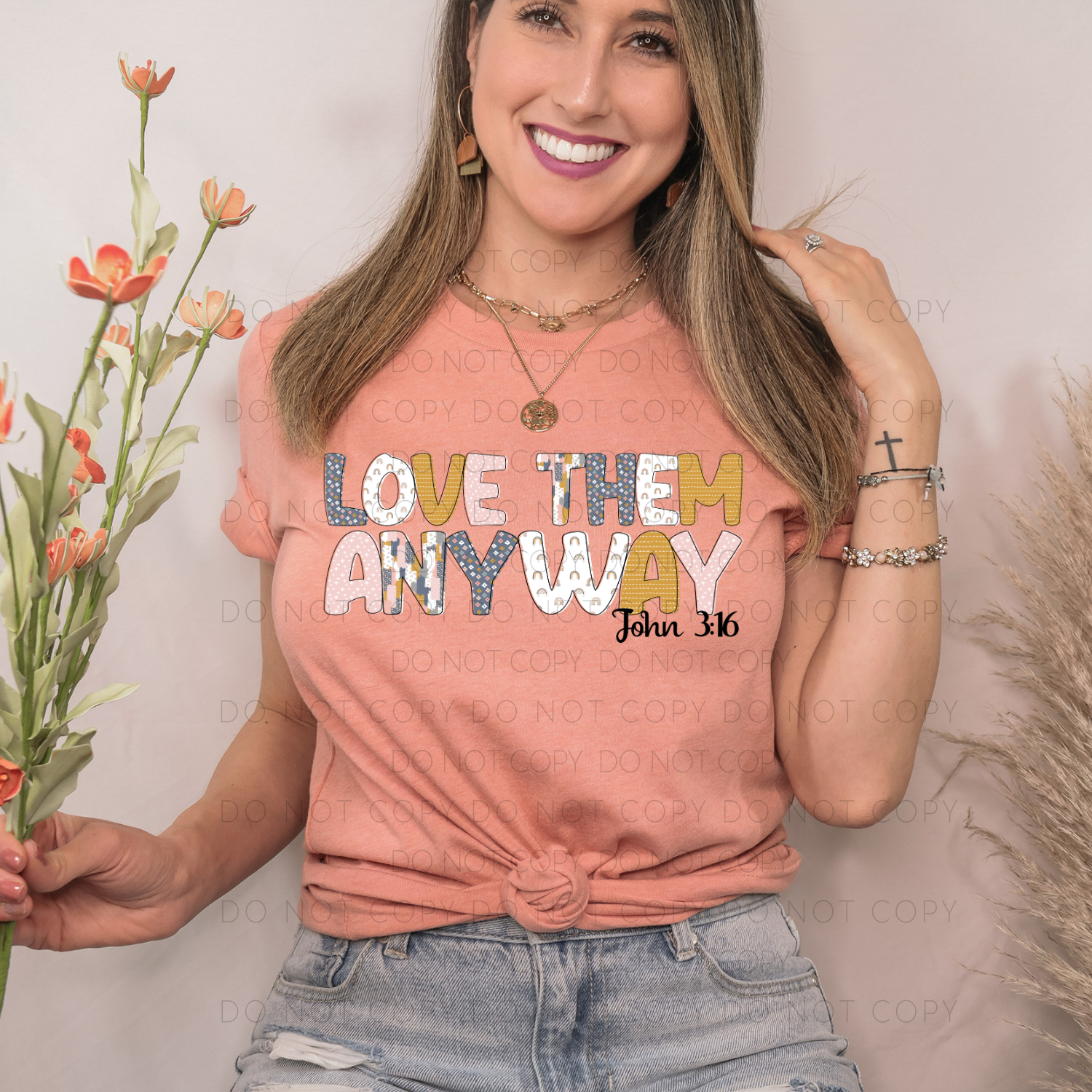 A person wearing a pink unisex t-shirt with the text 'LOVE THEM ANY WAY' printed on it, accompanied by a verse 'John 3:16'. The person is also wearing light blue jeans and accessorized with a necklace and a ring.