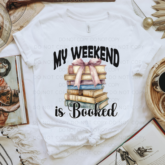 A white unisex T-shirt with the text 'MY WEEKEND IS BOOKED' printed on the front, styled with a graphic of stacked books and a bow.
